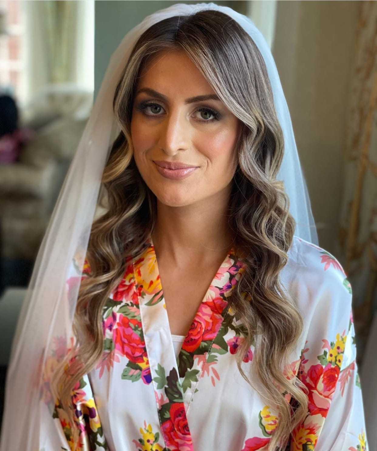 Bride in a floral robe with long curls and a veil, smiling softly indoors.
