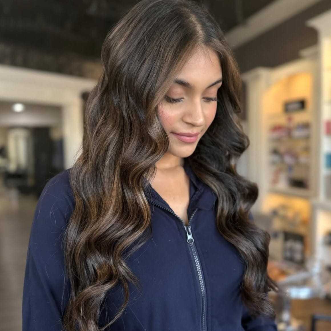 Young woman with long, wavy brown hair in a salon setting, smiling softly.