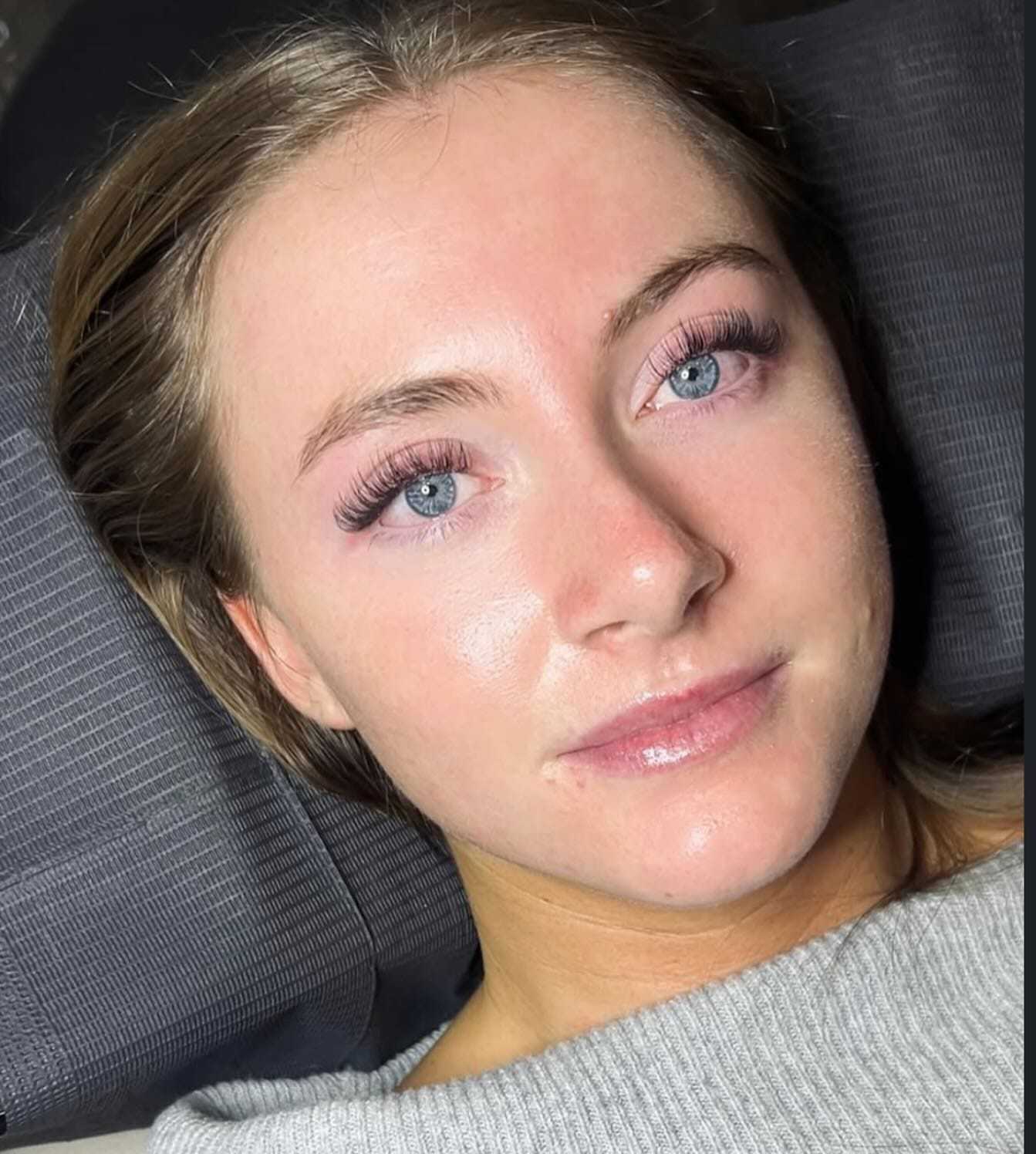 Woman with long eyelashes lying on a dark surface, wearing a light grey top.